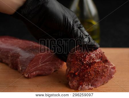 Hand In Black Glove Hold Beef Steak With Spices And Oil On A Wooden Board With A Black Background