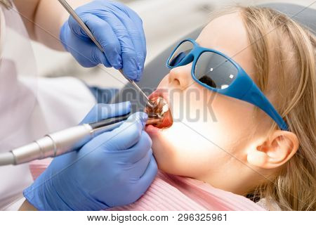 Dentist performing dental procedure to a little girl in pediatric dental clinic. Doctor using prophy brush on handpiece to polish filling. Calm child is sitting in a dental chair wearing sunglasses