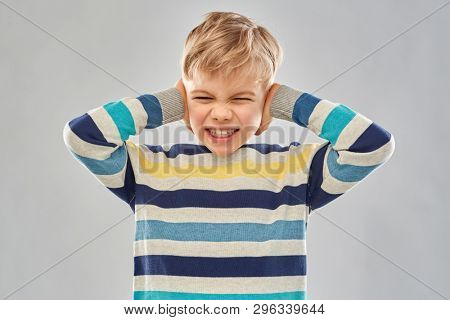 noise, stress and people concept - stressed little boy in striped pullover closing ears by hands over grey background