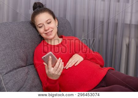 Indoor Shot Of Happy Pregnant Female Holding Smart Phone, Wearing Red Sweater And Leggins, Has Bunch