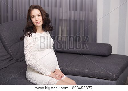 Beautiful Young Future Mom In Gentle White Dress, Has Dark Wavy Hair, Sits On Grey Couch Sofa At Hom