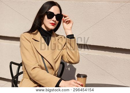 Black Haired European Magnetic Female Holding Brown Papercup In One Hand, Touching Black Sunglasses,