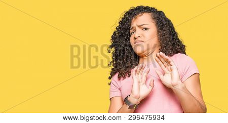 Young beautiful woman with curly hair wearing pink t-shirt disgusted expression, displeased and fearful doing disgust face because aversion reaction. With hands raised. Annoying concept.