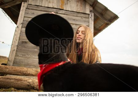 Happy Young Woman Plaing With Her Black Dog In Fron Of Old Wooden House. Girl Tries A Hat To Her Dog
