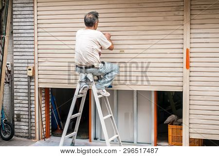 An Elderly Man Repairing Metal Blinds, Sitting On The Stairs . The View From The Back