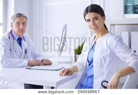 A Doctor Smiling At The Camera With Her Male Colleage In The Back Of The Consulting Room In Hospital
