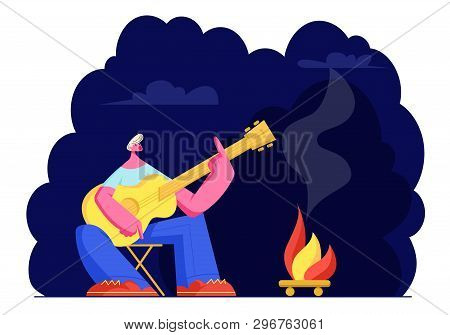 Young Man Sitting At Campfire At Night Time Singing Songs And Playing Guitar. Male Tourist Sparetime
