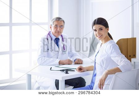 A doctor smiling at the camera with her male colleage in the back of the consulting room in hospital
