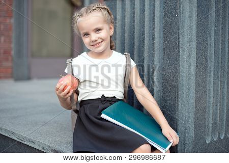 Happy Little Schoolgirl With Blond Hair In School Uniform With Lunch, Book Sits Near School.