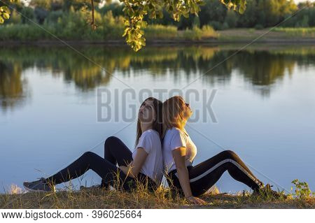 Two Athletic Young Women Are Resting After Training On The River Bank. Blonde And The Brunette Are B
