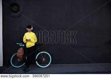Front view of a fashionable young mixed race transgender adult in the street, texting on the smartphone standing next to a bike with headphones on against a grey wall