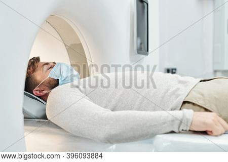Young male patient in casualwear and protective mask lying on long couchette and moving into large medical equipment for examination