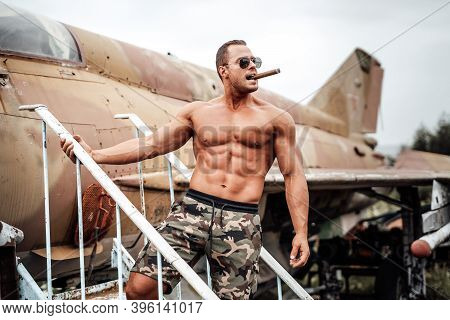 Portrait Of Muscular Naked Bodybuilder Smoking Cigar On Stairs Near Airplane In Daytime.