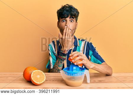 Young hispanic man sitting on the table using juicer covering mouth with hand, shocked and afraid for mistake. surprised expression 