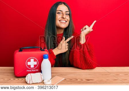 Beautiful hispanic woman with first aid kit smiling and looking at the camera pointing with two hands and fingers to the side. 