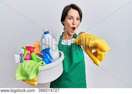 Young brunette woman with short hair wearing apron holding cleaning products pointing with finger surprised ahead, open mouth amazed expression, something on the front 
