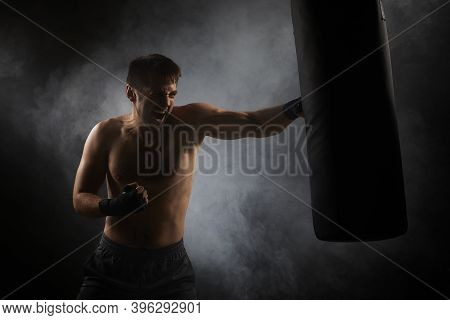 Aggressive Boxer In Black Boxing Wraps Punching In Boxing Bag On Dark Background With Smoke