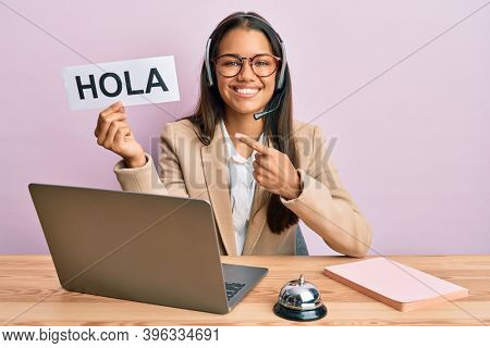 Beautiful hispanic woman wearing operator headset holding hola greenting smiling happy pointing with hand and finger 