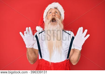 Old senior man wearing santa claus costume with suspenders angry and mad screaming frustrated and furious, shouting with anger looking up. 