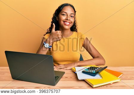 Young african american girl working at the office with laptop and calculator doing happy thumbs up gesture with hand. approving expression looking at the camera showing success. 