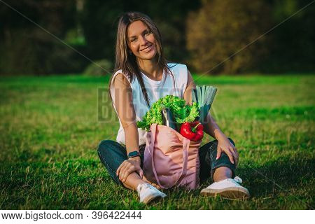 Beautiful Vegan Woman Sitting On The Grass With Pink Reusable Textile Shopping Bag With Fresh Bio Ve