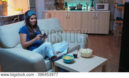 Woman Sitting On Sofa Playing Video Game Late At Night Wering Eye Mask On Forehead. Excited Determin