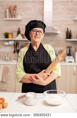 Professional Baker Smiling At Camera In Home Kitchen Wearing Apron And Bonete. Retired Elderly Baker