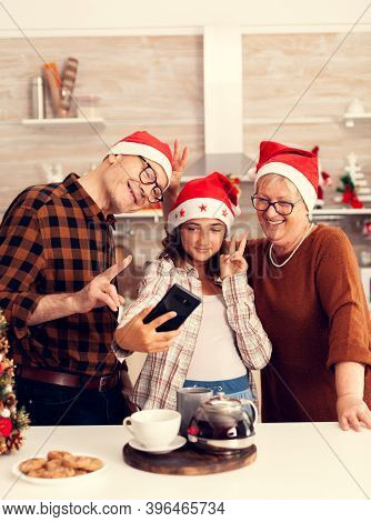 Niece Taking Selfie With Granddparents Celebrating Christmas In Decorated Kitchen. Happy Cheerful Mu