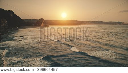 Rock ocean shore sunset with waves at water surface in aerial view. Epic nobody nature scenery of sun setting over wavy seascape. Summer sunlight at cliff coastline. Rocky silhouette at sunny evening