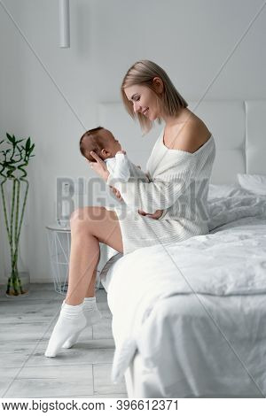 Caucasian Woman Sitting On Bed With A Baby Who Has Just Woken Up. A Portrait Of A Mother Who Looks T