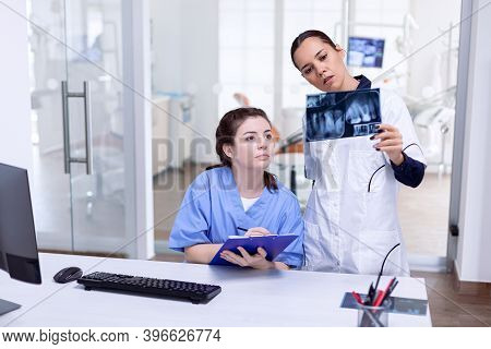 Dentist Holding Teeth X-ray Explaning Diagnosis For Nurse At Reception. Stomatolog And Her Assistant