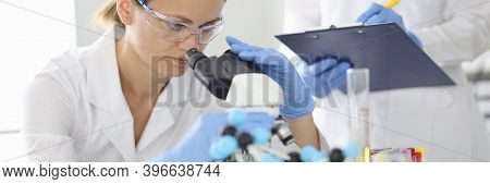 Woman Scientist In Protective Glasses Looks Through Microscope And Trains Assistant In Chemical Labo