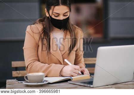 Young Female In Black Face Mask Sitting On Terrace In Cafe. Business Woman Working Outdoos In Quaran