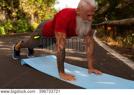 Senior Man With White Beard Doing Exercises On The Nature, Connecting Via Laptop, New Normal And Soc