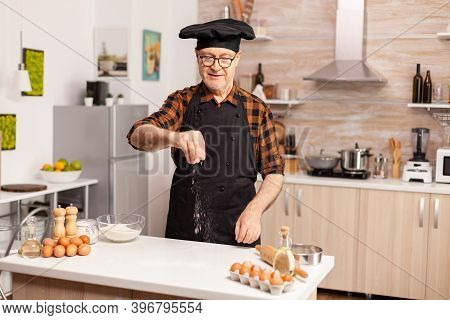 Senior Baker Sprinkling Whear Flour On Home Kitche Table For Tasty Recipe. Retired Chef With Bonete 