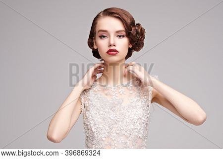 Evening makeup and hairstyle. Portrait of a charming young woman in a luxury white dress on a gray background. Hair and makeup in the style of the 20s. 