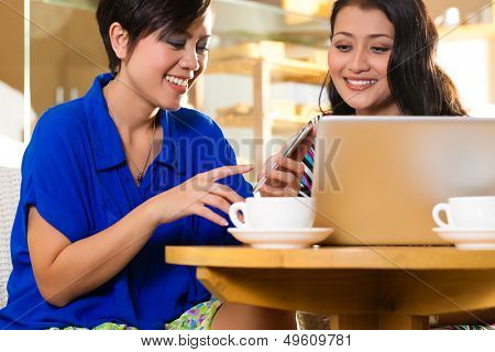 Asian female friends enjoying her leisure time in a cafe, drinking coffee or cappuccino and working on a laptop computer