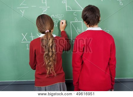 Rear view of teenage girl writing on board with classmate standing in classroom