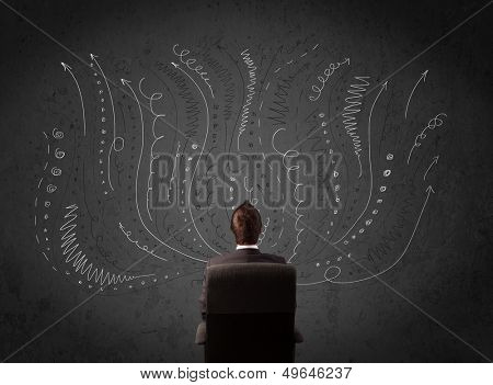 Thoughtful young businessman sitting and deciding in front of a chalkboard with sketched arrows and lines in different directions