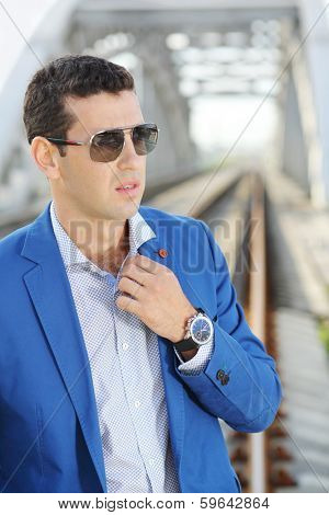 Handsome man in blue jacket and sunglasses stands on railroad bridge.