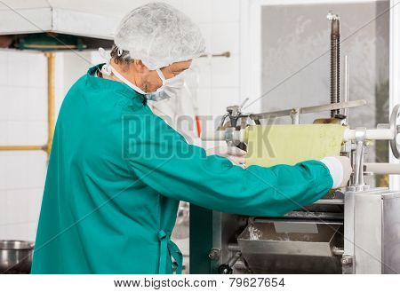 Side view of male chef in protective workwear adjusting pasta sheet into machine at commercial kitchen