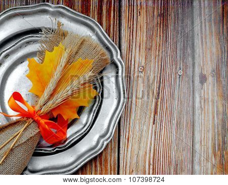 A rustic place setting with maple leaf and wheat decorations on rough wooden background. Old pewter plates, stained silver-plated flatware with a burlap napkin tied with a bright orange ribbon.