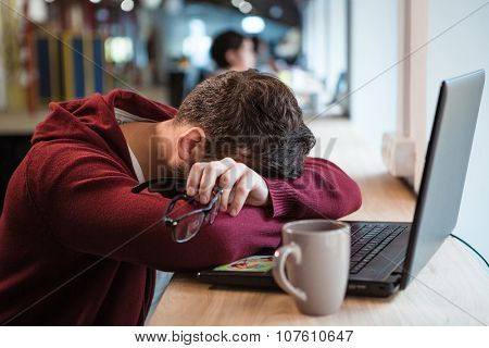 Tired man in brown sweetshirt holding glasses while sleeping at office desk
