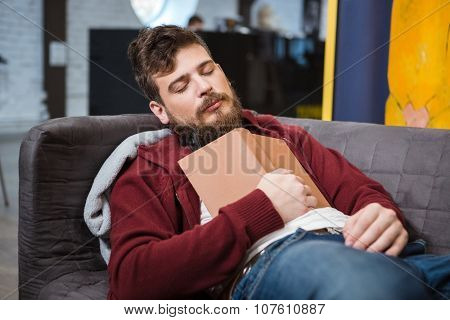 Bearded handsome tired man sleeping on grey sofa holding book