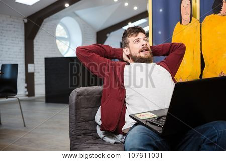 Handsome tired guy with beard yawning while working on laptop at office sitting on grey sofa
