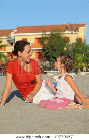 Мать с дочерью сидеть на песок и смех вблизи двухэтажных коттеджах