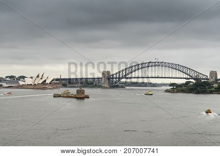 Sydney Australia - March 21 2017: Sydney Harbour bridge Opera House and Denison Fort under gray rainy sky. Darker gray water in front with small boats drawing white lines.