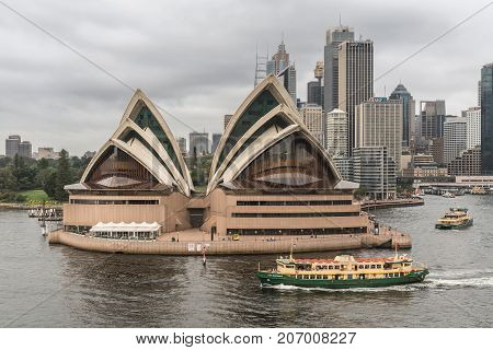 Sydney Australia - March 21 2017: Frontal view of Opera House under gray rainy sky. City skyline in back. Ferry boats on the water in front people on the platforms.