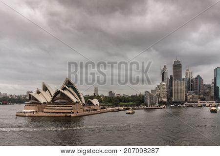 Sydney Australia - March 21 2017: Frontal view of Opera House under brown-gray rainy sky. Part of City skyline in back. Ferry boats on the water people on the platforms.