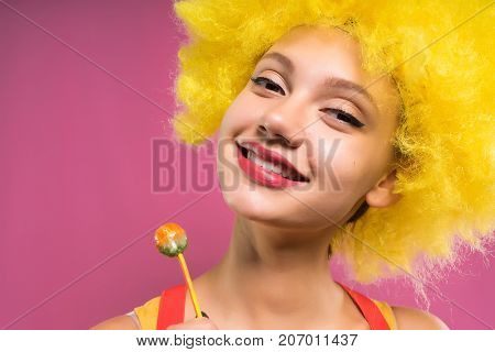 laughing girl in a bright orange wig on a pink background holds in hands chupa chups